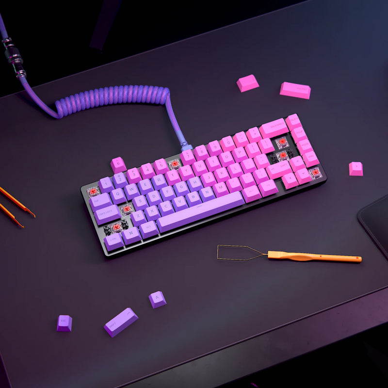 A Black GMMK 2 65% with GPBT Nebula keycaps is shown on a desk with some keycaps removed, exposing the switches underneath. A black wrist rest is positioned in front of the keyboard, and a Nebula coiled cable is connected to it. Additional keycaps and tools are scattered around the desk.